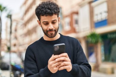 Genç Arap adam sokakta akıllı telefon kullanarak kendinden emin gülümsüyor.
