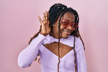 African woman with braided hair standing over pink background smiling with hand over ear listening an hearing to rumor or gossip. deafness concept. 