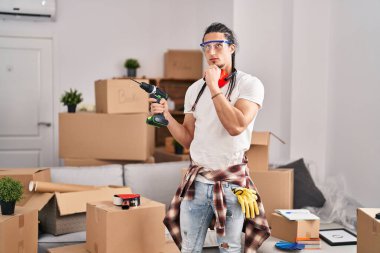 Hispanic man with long hair holding screwdriver at new home serious face thinking about question with hand on chin, thoughtful about confusing idea 