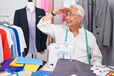 Middle age man with grey hair dressmaker using sewing machine very happy and smiling looking far away with hand over head. searching concept. 