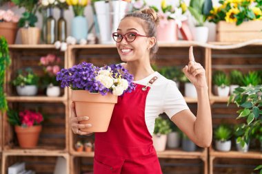 Young hispanic woman working at florist shop holding plant smiling with an idea or question pointing finger with happy face, number one 