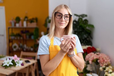 Young blonde woman florist smiling confident drinking cup of coffee at flower shop