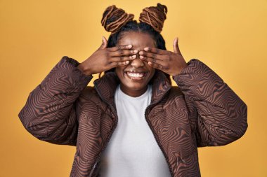 African woman with braided hair standing over yellow background covering eyes with hands smiling cheerful and funny. blind concept. 