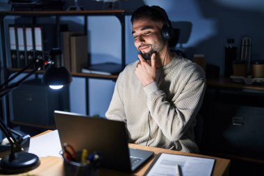 Young handsome man working using computer laptop at night looking confident at the camera smiling with crossed arms and hand raised on chin. thinking positive. 