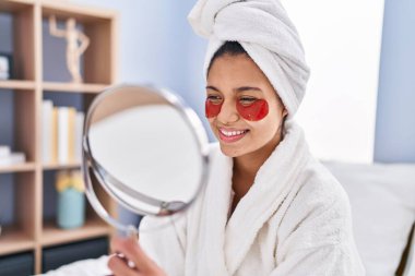 Yatak odasındaki aynaya bakan genç Latin kadın dolgun göz tedavisi görüyor.