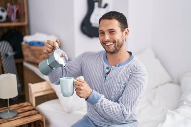 Young man drinking cup of coffee sitting on bed at bedroom