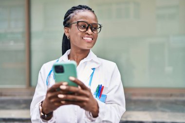 Afro-Amerikalı kadın doktor hastanede akıllı telefon kullanarak gülümsüyor.