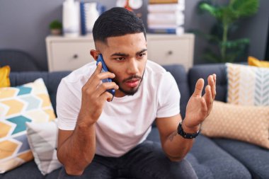 Young latin man talking on the smartphone sitting on sofa at home