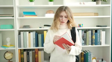 Young blonde woman student reading book standing at library university