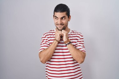 Hispanic man with long hair standing over isolated background laughing nervous and excited with hands on chin looking to the side 