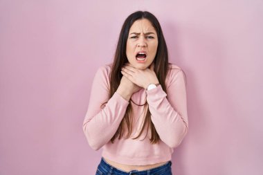 Young brunette woman standing over pink background shouting suffocate because painful strangle. health problem. asphyxiate and suicide concept. 