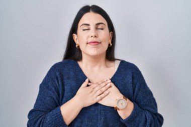 Genç esmer kadın izole edilmiş arka planda duruyor. Elleri göğsünde, gözleri kapalı gülümsüyor ve yüzünde minnet dolu bir jest var. sağlık kavramı. 