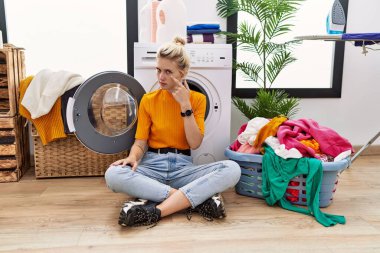 Young blonde woman doing laundry sitting by washing machine pointing to the eye watching you gesture, suspicious expression 