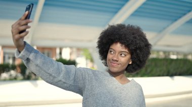 Afro-Amerikalı kadın, parkta akıllı telefonun yanında kendinden emin bir şekilde selfie çekiyor.