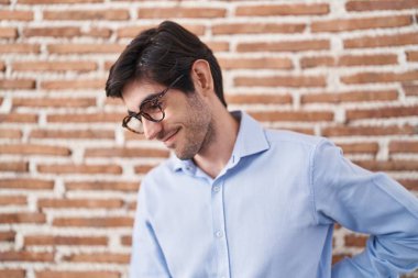 Young hispanic man standing over brick wall background suffering of backache, touching back with hand, muscular pain 