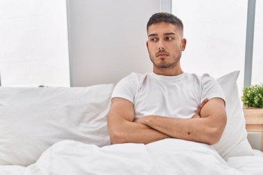Young hispanic man with serious expression sitting on bed at bedroom