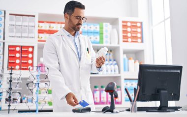 Young hispanic man pharmacist holding pills using credit card and dataphone at pharmacy