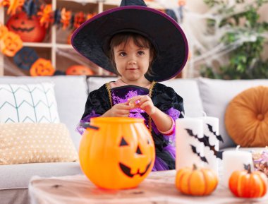 Adorable hispanic girl having halloween party putting sweets on pumpkin basket at home