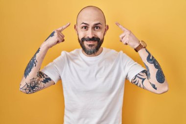 Young hispanic man with tattoos standing over yellow background smiling pointing to head with both hands finger, great idea or thought, good memory 