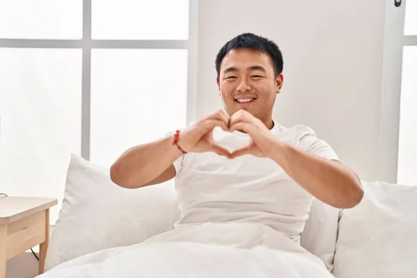 Young chinese man doing heart gesture with hands sitting on bed at bedroom