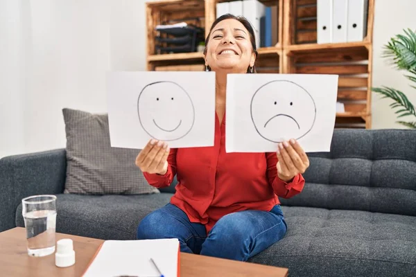 Middle age hispanic woman working on depression holding sad to happy emotion paper smiling and laughing hard out loud because funny crazy joke.