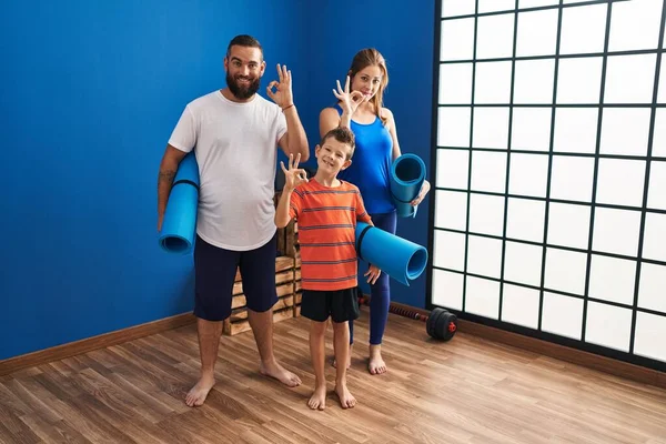 Family of three holding yoga mat doing ok sign with fingers, smiling friendly gesturing excellent symbol