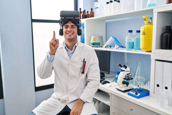 Young hispanic man working at scientist laboratory wearing vr glasses smiling with an idea or question pointing finger up with happy face, number one