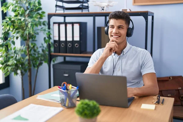 Young hispanic man working at the office wearing headphones looking confident at the camera smiling with crossed arms and hand raised on chin. thinking positive. 