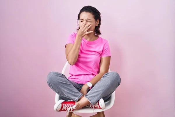 ヒスパニック系の若い女性がピンクの背景の上に椅子に座って何か臭くて嫌な 耐え難い匂いを嗅ぎ 鼻の上に指で息を保持する 臭いが悪い — ストック写真
