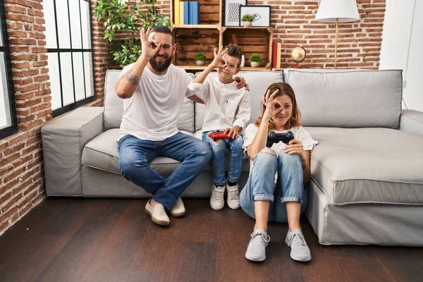 Family of three playing video game sitting on the sofa smiling happy doing ok sign with hand on eye looking through fingers