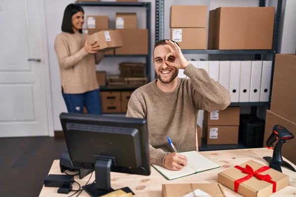 Two young people working at small business ecommerce smiling happy doing ok sign with hand on eye looking through fingers