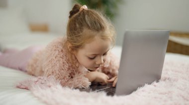 Adorable blonde girl using laptop lying on bed at bedroom