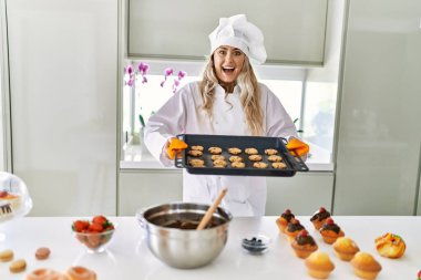 Young woman wearing cook uniform holding oven tray with cookies at kitchen