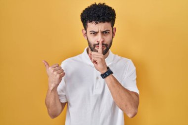 Arab man standing over yellow background asking to be quiet with finger on lips pointing with hand to the side. silence and secret concept. 