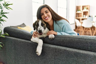 Evde köpekle koltukta otururken kendinden emin gülümseyen genç kadın.