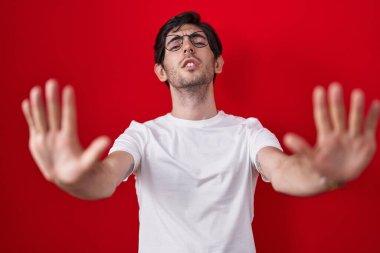 Young hispanic man standing over red background doing stop gesture with hands palms, angry and frustration expression 