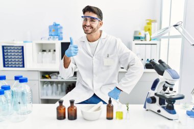 Laboratuvarda çalışan Latin asıllı genç adam mutlu ve pozitif gülümsüyor, başparmağı mükemmel ve onay işareti yapıyor. 