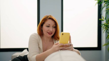 Genç kızıl saçlı kadın akıllı telefon kullanıyor. Evdeki koltukta oturuyor.