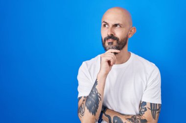 Hispanic man with tattoos standing over blue background with hand on chin thinking about question, pensive expression. smiling and thoughtful face. doubt concept. 