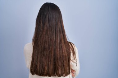 Genç esmer kadın mavi arka planda dikiliyor geriye bakıyor ve kollarını kavuşturuyor. 