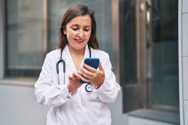 Middle age woman wearing doctor uniform using smartphone at street