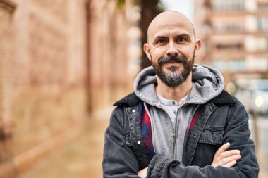 Kel genç adam kollarını kavuşturmuş sokakta el kol hareketi yapıyor.