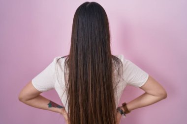 Chinese young woman standing over pink background standing backwards looking away with arms on body 