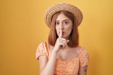 Young redhead woman standing over yellow background wearing summer hat asking to be quiet with finger on lips. silence and secret concept. 