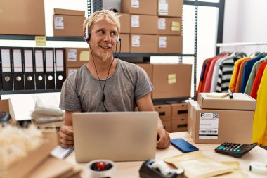Young blond man wearing operator headset working at online shop looking away to side with smile on face, natural expression. laughing confident. 