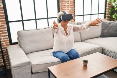 Evde sanal gerçeklik gözlüğü kullanan orta yaşlı bir kadın video oyunu oynuyor.