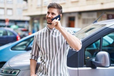 Beyaz bir adam akıllı telefondan konuşuyor. Sokakta arabaya yaslanıyor.