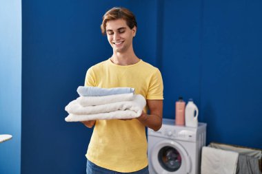 Young caucasian man smiling confident holding folded towels at laundry room