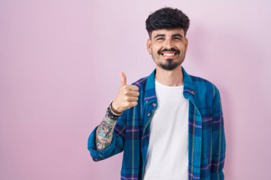 Pembe arka planda sakallı genç İspanyol adam elleriyle mutlu bir jest yapıyor. Kameraya bakıp başarıyı gösteren ifade. 