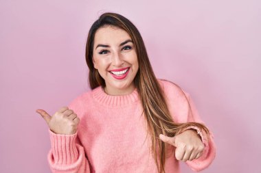 Pembe arka planda duran genç İspanyol kadın, arka tarafı işaret ediyor, baş parmakları havada, kendinden emin gülümsüyor. 